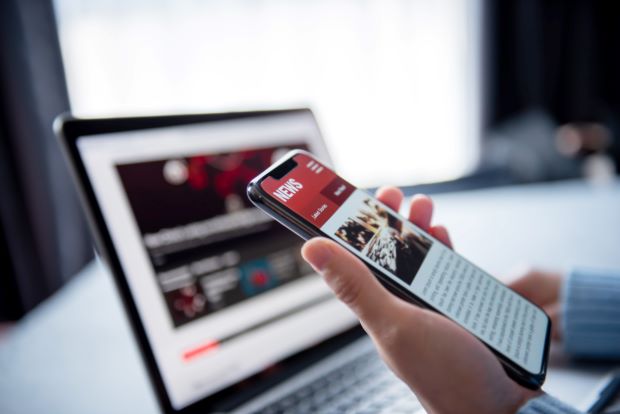 Close-up of someone looking at a news site on a smartphone with a laptop in the background