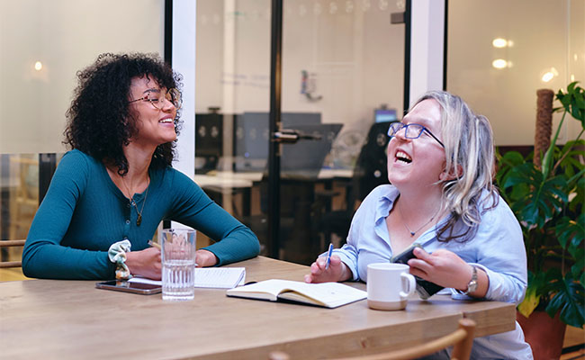 Two women laughing and writing in notebooks in an open office. 