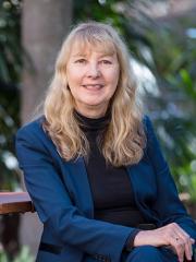 Professor Emerita Jennifer Corrin sitting on a chair in front of a garden.