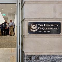 View of UQ Brisbane city with a plaque on the stone pillar and people inside socialising. 