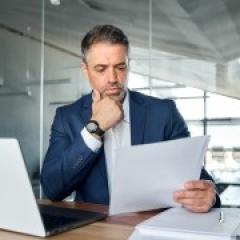 Serious busy mid aged professional business man wearing suit holding corporate documents reading paper contract sitting at desk in office.