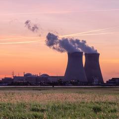 subject is a nuclear powerplant in the distance at sunset. There are two giant chimneys with smoke coming out of them. 