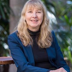 Professor Emerita Jennifer Corrin sitting on a chair in front of a garden.
